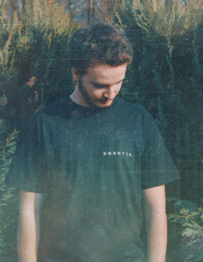 Borrtex is standing in front of a small trees and looking down. He is wearing a T-Shirt of his own brand with Borrtex logo.