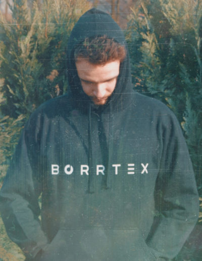 Borrtex is standing in front of a small trees and looking down. He is wearing a jacket of his own brand with Borrtex logo.