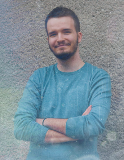 A close up photo of Borrtex with a blue sweater as he is standing next to a wall with hands crossed and smiling.