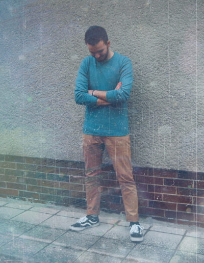 A photo of Borrtex with a blue sweater as he is standing next to a wall with hands crossed and smiling. He is looking down.
